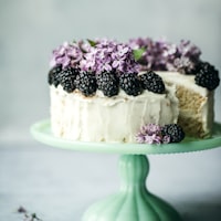 A white iced cake topped with blackberries and purple flowers on a green cake stand