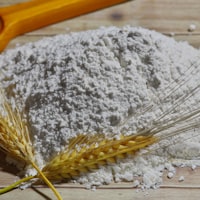 A heap of white flour poured from a yellow scoop, with ears of wheat beside it