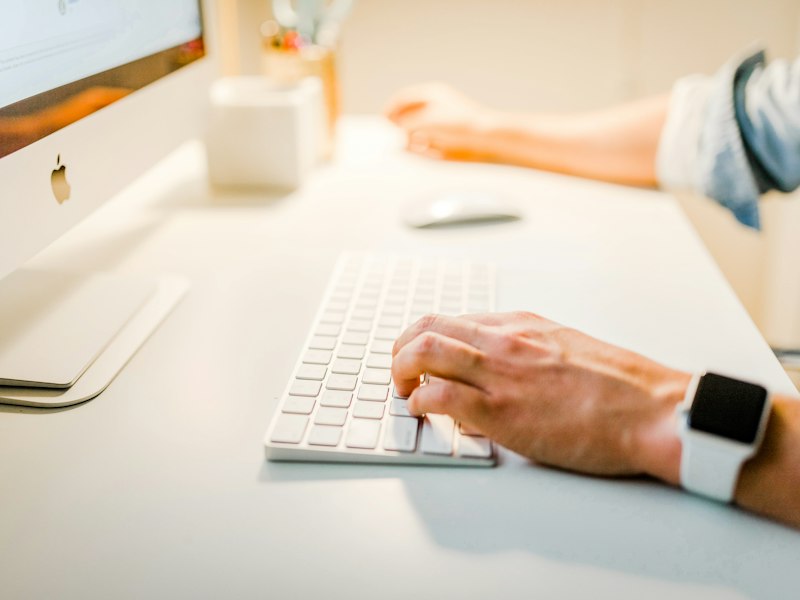 Hands typing on a keyboard at a bright desk beside a desktop monitor